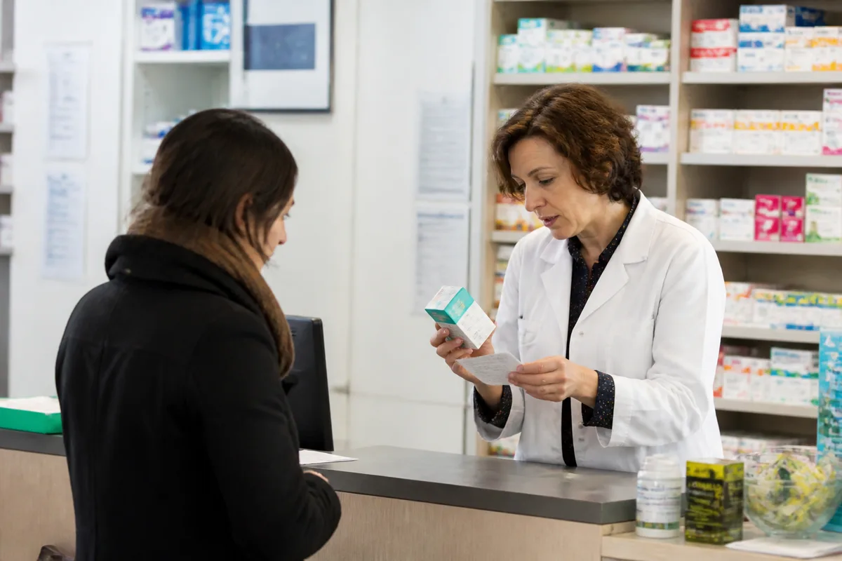 Pharmacien français vérifiant une boîte de médicament et une notice avec un patient au comptoir